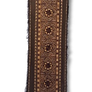 K&ouml;nnte beinhalten: Eine zylindrische Holzkiste mit einem dekorativen Muster aus Sternen und geometrischen Formen. Die Kiste ist aus hellem und dunklem Holz gefertigt und hat einen Fransenbesatz an der Ober- und Unterseite.