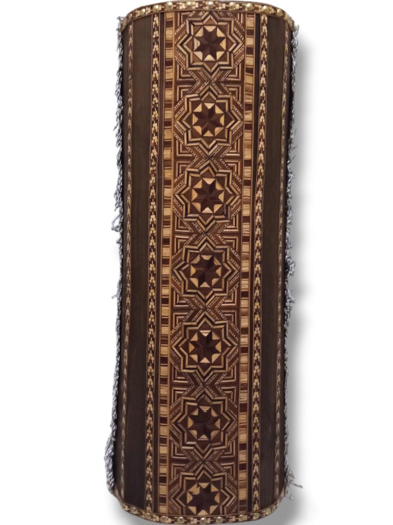 K&ouml;nnte beinhalten: Eine zylindrische Holzkiste mit einem dekorativen Muster aus Sternen und geometrischen Formen. Die Kiste ist aus hellem und dunklem Holz gefertigt und hat einen Fransenbesatz an der Ober- und Unterseite.