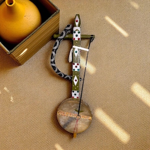 Pode incluir: Um instrumento de cordas decorativo e artesanal com um corpo redondo e um braço longo e decorado. O instrumento apresenta um design pintado em verde e branco com detalhes coloridos. Uma peça curva em preto e branco está presa ao braço. Um vaso amarelo está em uma caixa de madeira no fundo.