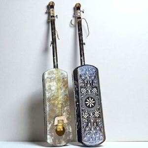 May include: Two handmade stringed instruments with long necks and rectangular bodies. One is aged with a light, textured finish, while the other is dark with intricate white floral patterns. Both have tuning pegs and strings.