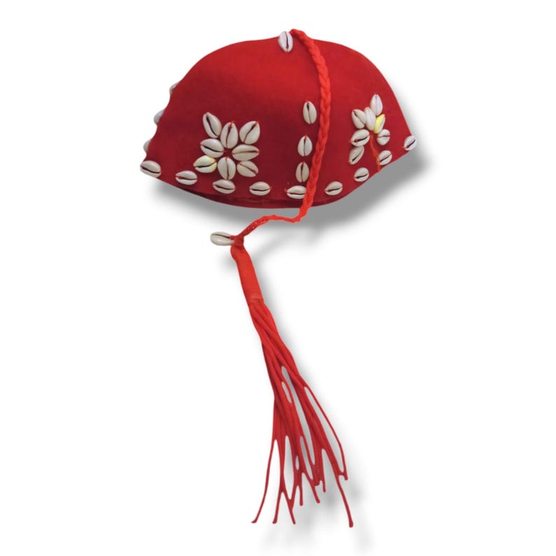 K&ouml;nnte beinhalten: Ein roter Filzhut mit einem wei&szlig;en Muschel-Design und einem langen roten Quasten.