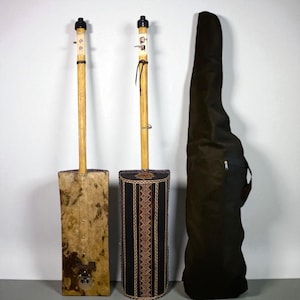 Puede incluir: Dos instrumentos de cuerda africanos hechos a mano, cada uno con un mástil largo de madera y un cuerpo rectangular o cilíndrico. Uno tiene un cuerpo de tela estampada, el otro un cuerpo de madera texturizada. También hay una funda de transporte negra.