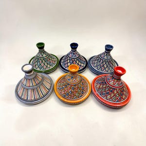 Puede incluir: Seis coloridas ollas de tagine de cerámica con tapas cónicas. Cada olla presenta un patrón geométrico único en varios colores, incluyendo verde, azul, naranja y rojo. Las ollas están dispuestas sobre una superficie blanca, mostrando sus diseños decorativos.