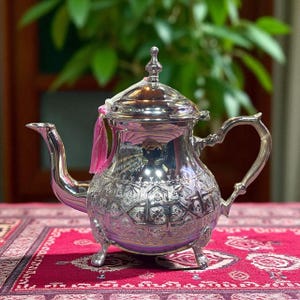 Peut inclure: Une théière argentée avec des motifs en relief complexes et un bec verseur incurvé. La théière a un couvercle avec un fleuron décoratif et une poignée au design unique. Un ruban rose est attaché. La théière est sur une surface rouge à motifs.