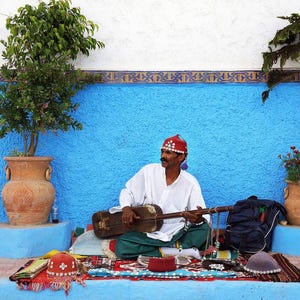 K&ouml;nnte beinhalten: Ein Mann mit einem rot-wei&szlig;en Hut spielt ein traditionelles Saiteninstrument vor einer blauen Wand. Er sitzt auf einem Teppich mit blau-wei&szlig;em Muster. Im Hintergrund befinden sich zwei Topfpflanzen.