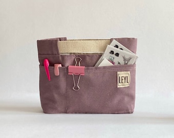 May include: A mauve canvas organizer with multiple pockets, holding a pink pen, a small pink object, a pink binder clip, and stickers. The organizer has a beige interior and a small label that reads "LEYL".