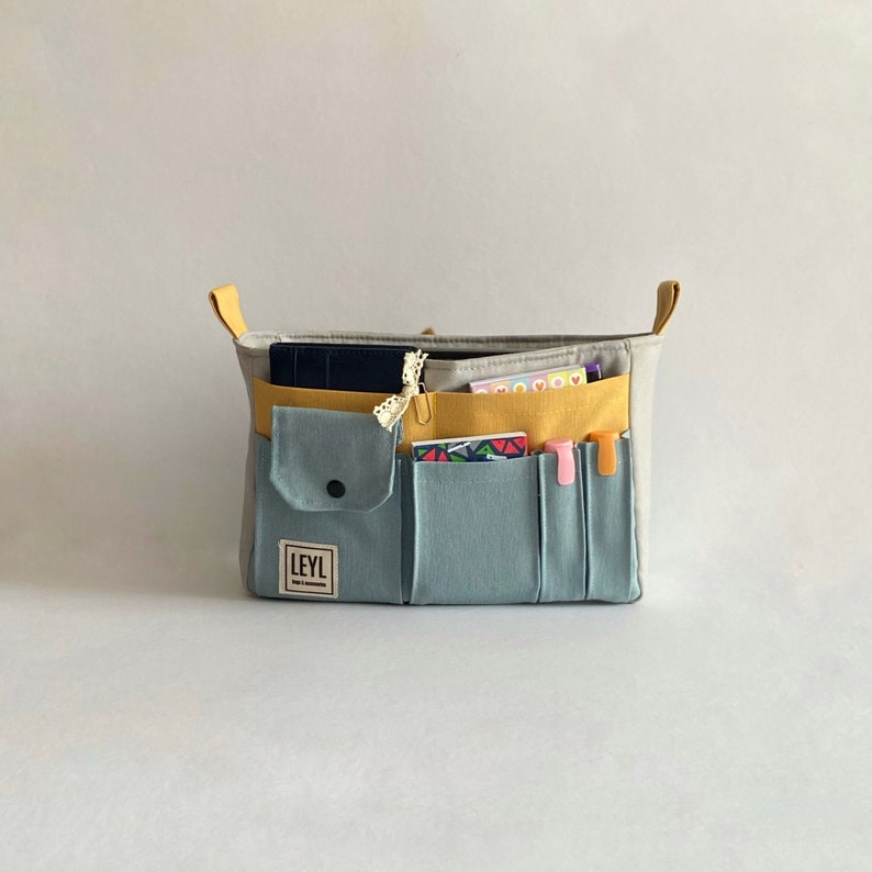 May include: A gray and blue canvas tote bag with multiple pockets and a yellow zippered compartment. The bag is filled with various items, including a notebook, pens, and a small notepad. The bag has the word "LEYL" printed on the front.