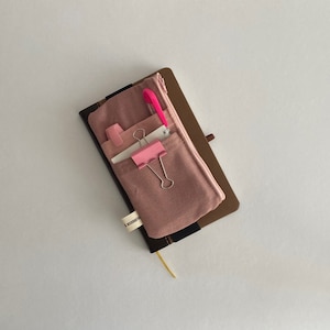 Könnte beinhalten: Eine rosa Stofftasche mit Reißverschluss, die einen rosa Stift, eine rosa Büroklammer und eine weiße Karte enthält. Die Tasche ist an einem braunen Ledernotizbuch befestigt.