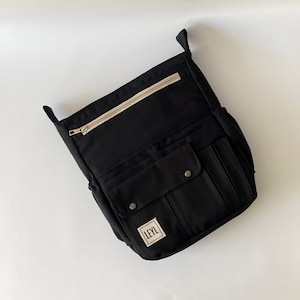 May include: Black messenger bag with multiple pockets and a beige zipper. The bag has a rectangular label with the text "LEYL". The bag has two handles and two side pockets.