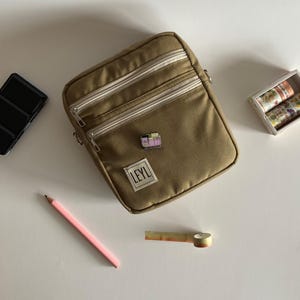 Op de afbeelding: Kleurige crossbodytas met drie vakken met ritssluiting en een kleine speld. De tas heeft een label met de tekst "LEYL". Een roze potlood, aquarelpalet en decoratieve tapetrollen zijn ook zichtbaar.