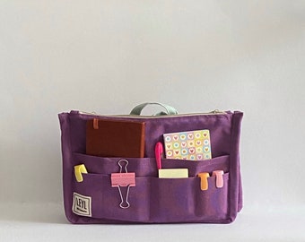 May include: A purple organizer bag with multiple pockets, holding a brown notebook, a pink pen, and a notepad with heart designs. The bag also contains yellow and pink binder clips. The bag has a zipper closure and a light green handle.