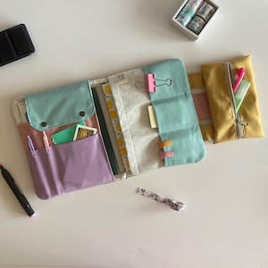 Könnte beinhalten: Ein offener Organizer mit mehreren Taschen in Pastellfarben. Der Organizer hat eine hellblaue Klappe mit zwei Knöpfen, eine lila Tasche und eine gelbe Reißverschlusstasche. Im Inneren befinden sich Stifte, Karten und ein Notizbuch. Ein schwarzer Marker und dekoratives Klebeband sind ebenfalls sichtbar.