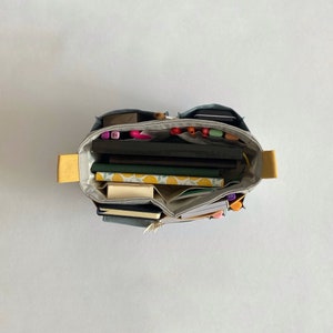 May include: A gray canvas tote bag with yellow handles is filled with various items, including notebooks, pens, and markers. The bag is open and the contents are visible.