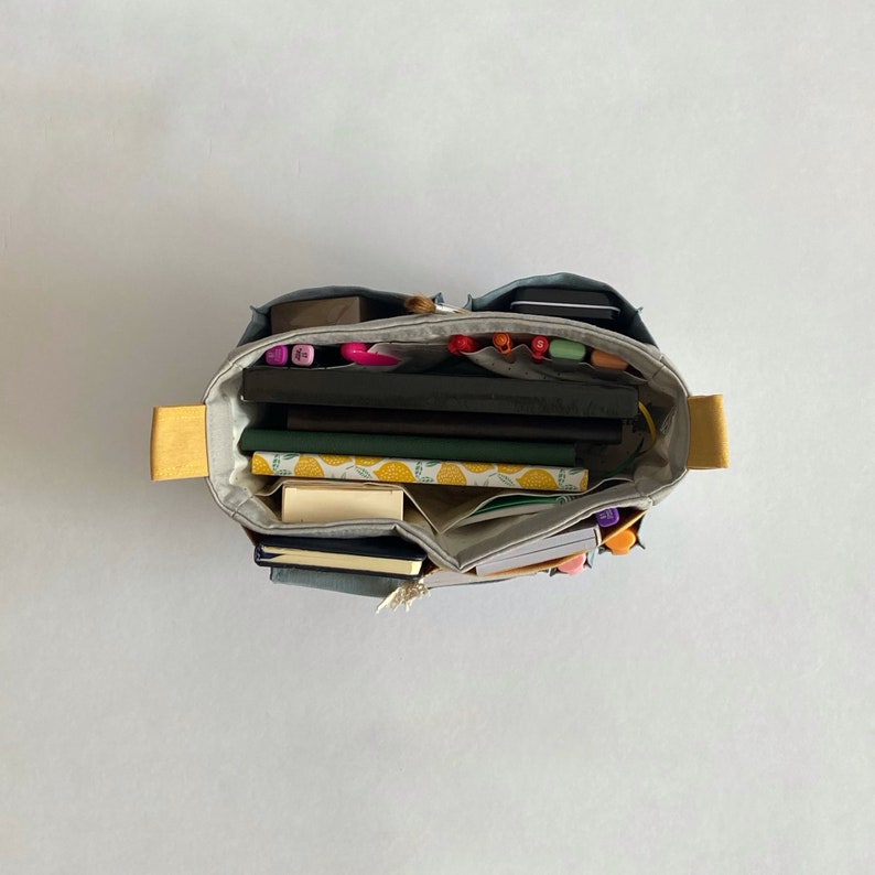 May include: A gray canvas tote bag with yellow handles is filled with various items, including notebooks, pens, and markers. The bag is open and the contents are visible.