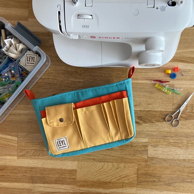 May include: A turquoise and yellow fabric sewing organizer with multiple pockets. The organizer has a label that says "LEYL" and a small button on the front.