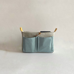 May include: A light blue organizer with two pockets and a beige fabric exterior. The organizer has yellow handles and a paintbrush is visible inside.