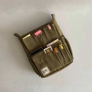 May include: An olive green canvas organizer with multiple pockets and a label that reads "LEYL". The organizer contains various items, including paper clips, sticky notes, and a small photo. It has two handles at the top.