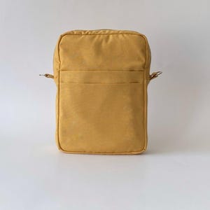 May include: Mustard-yellow crossbody bag with a rectangular shape. The bag features a front pocket and metal rings for a shoulder strap. The material has a slightly textured appearance, and the bag is set against a white background.
