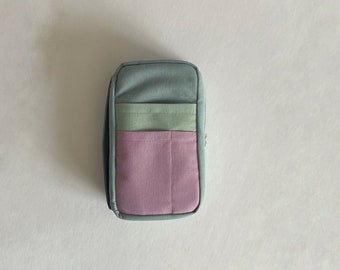 May include: A rectangular fabric pencil case featuring a color-blocked design. The main body is a muted blue-gray, accented by a horizontal light sage green pocket and a larger, vertical-stitched dusty rose pink pocket on the front.