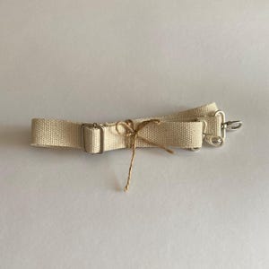 May include: A beige canvas bag strap featuring a silver-toned metal adjustable slider and two silver-toned swivel lobster clasps at the ends. A thin brown twine string is tied in a small bow around the center of the woven fabric strap.