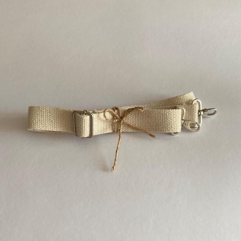 May include: A beige canvas bag strap featuring a silver-toned metal adjustable slider and two silver-toned swivel lobster clasps at the ends. A thin brown twine string is tied in a small bow around the center of the woven fabric strap.