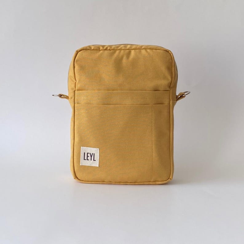 May include: Mustard yellow crossbody bag with a rectangular shape. The bag has a front pocket and a small white label with the text "LEYL". The bag has silver metal rings for the strap.