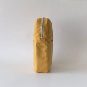 May include: Mustard-yellow tote bag with a rectangular shape. The bag has a silver zipper running down the center and two small pockets on the front. The bag is made of a textured fabric and is designed to be carried by hand.