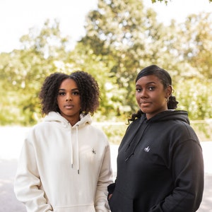 May include: Two women wearing hoodies. The woman on the left is wearing a white hoodie with a small logo on the front. The woman on the right is wearing a black hoodie with a small logo on the front.
