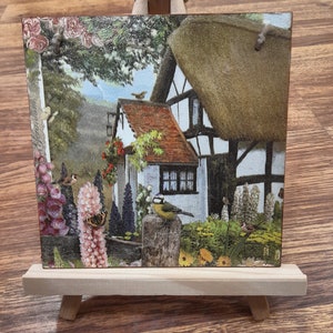 May include: A small wooden easel displaying a square tile with a colorful image of a cottage with a thatched roof, surrounded by flowers and birds. The cottage is white with brown trim and has a red tiled roof. The image is set against a green background with a blue sky.