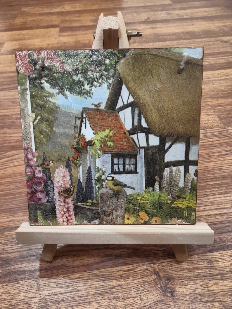 May include: A small wooden easel displaying a square tile with a colorful image of a cottage with a thatched roof, surrounded by flowers and birds. The cottage is white with brown trim and has a red tiled roof. The image is set against a green background with a blue sky.
