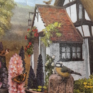 May include: A quaint cottage with a brown tiled roof and white walls, surrounded by lush greenery. A small bird perches on the roof, while another bird sits on a wooden post in the garden. The garden features a variety of flowers, including pink lupines and white roses.