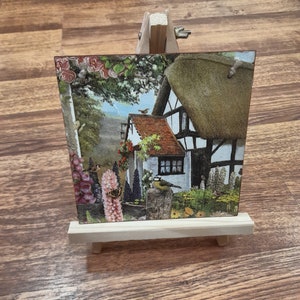 May include: A small wooden easel with a postcard featuring a cottage with a thatched roof, surrounded by flowers and birds. The postcard is printed with a vintage aesthetic.