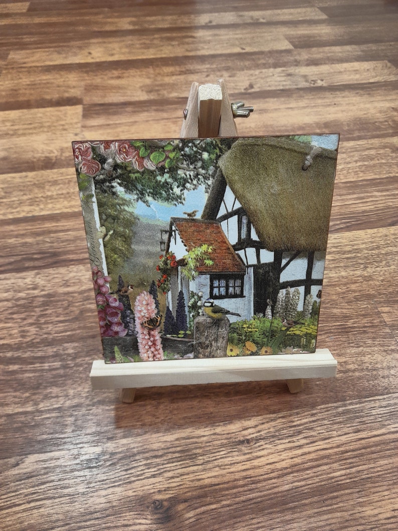 May include: A small wooden easel with a postcard featuring a cottage with a thatched roof, surrounded by flowers and birds. The postcard is printed with a vintage aesthetic.