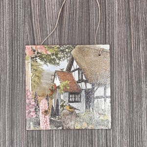 May include: A decorative wooden plaque with a printed image of a quaint cottage with a thatched roof, surrounded by lush greenery and flowers. A small bird is perched on a branch near the cottage.
