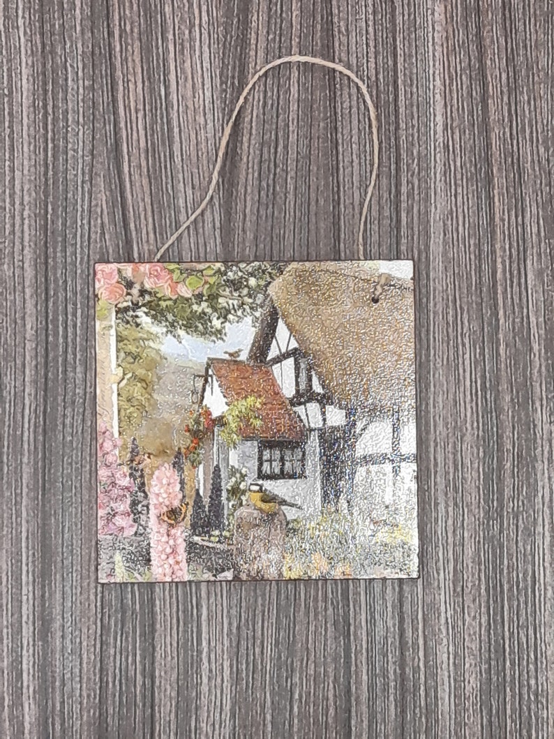 May include: A decorative wooden plaque with a printed image of a quaint cottage with a thatched roof, surrounded by lush greenery and flowers. A small bird is perched on a branch near the cottage.