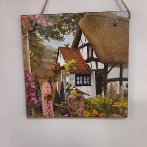 May include: A wooden wall hanging with a colorful image of a cottage with a thatched roof, surrounded by flowers and birds. The cottage is white with black trim and has a red tile roof. The flowers are in shades of pink, purple, and yellow. The birds are brown and yellow.