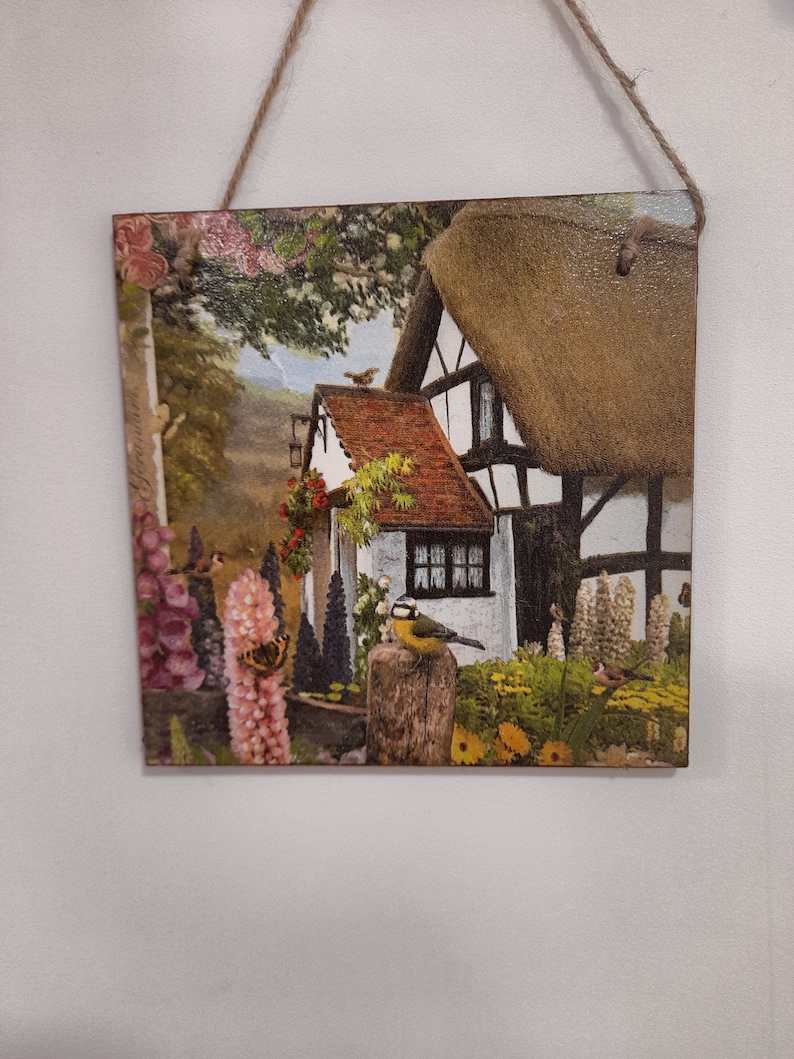 May include: A wooden wall hanging with a colorful image of a cottage with a thatched roof, surrounded by flowers and birds. The cottage is white with black trim and has a red tile roof. The flowers are in shades of pink, purple, and yellow. The birds are brown and yellow.