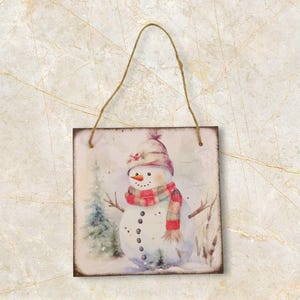 K&ouml;nnte beinhalten: Quadratisches Holzschild mit einer Aquarellillustration eines Schneemanns mit Hut und Schal. Der Schneemann hat eine Karottennase und Knopfaugen. Eine Schnurschlaufe zum Aufh&auml;ngen ist angebracht. Der Hintergrund zeigt eine verschneite Winterlandschaft.