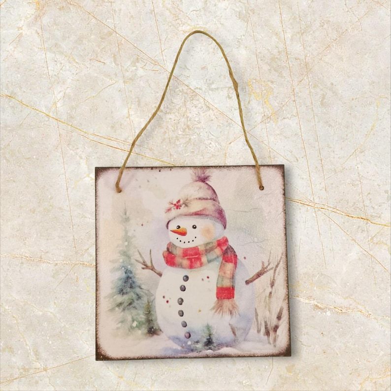 K&ouml;nnte beinhalten: Quadratisches Holzschild mit einer Aquarellillustration eines Schneemanns mit Hut und Schal. Der Schneemann hat eine Karottennase und Knopfaugen. Eine Schnurschlaufe zum Aufh&auml;ngen ist angebracht. Der Hintergrund zeigt eine verschneite Winterlandschaft.