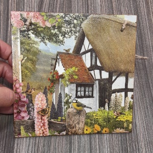 May include: A vintage-style illustration of a cottage with a thatched roof, surrounded by a lush garden with flowers, birds, and butterflies. The cottage has a white exterior with black trim and a red tiled roof.