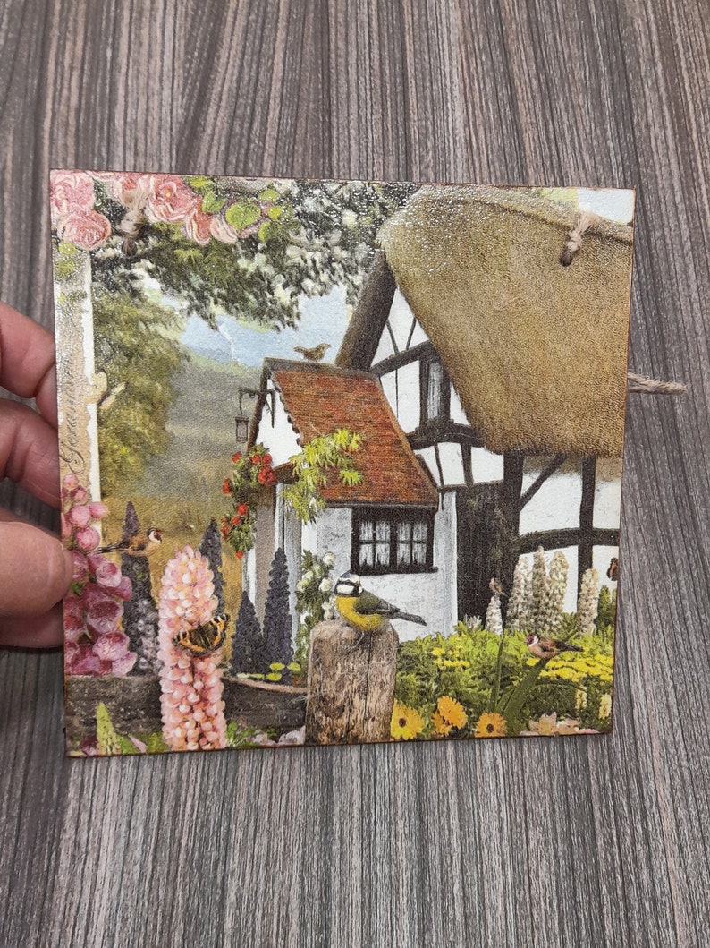 May include: A vintage-style illustration of a cottage with a thatched roof, surrounded by a lush garden with flowers, birds, and butterflies. The cottage has a white exterior with black trim and a red tiled roof.