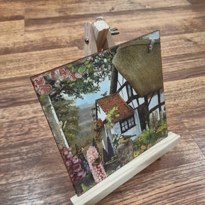 May include: A wooden easel with a small painting of a cottage with a thatched roof and flowers. The painting is on a wooden board and the easel is made of natural wood.