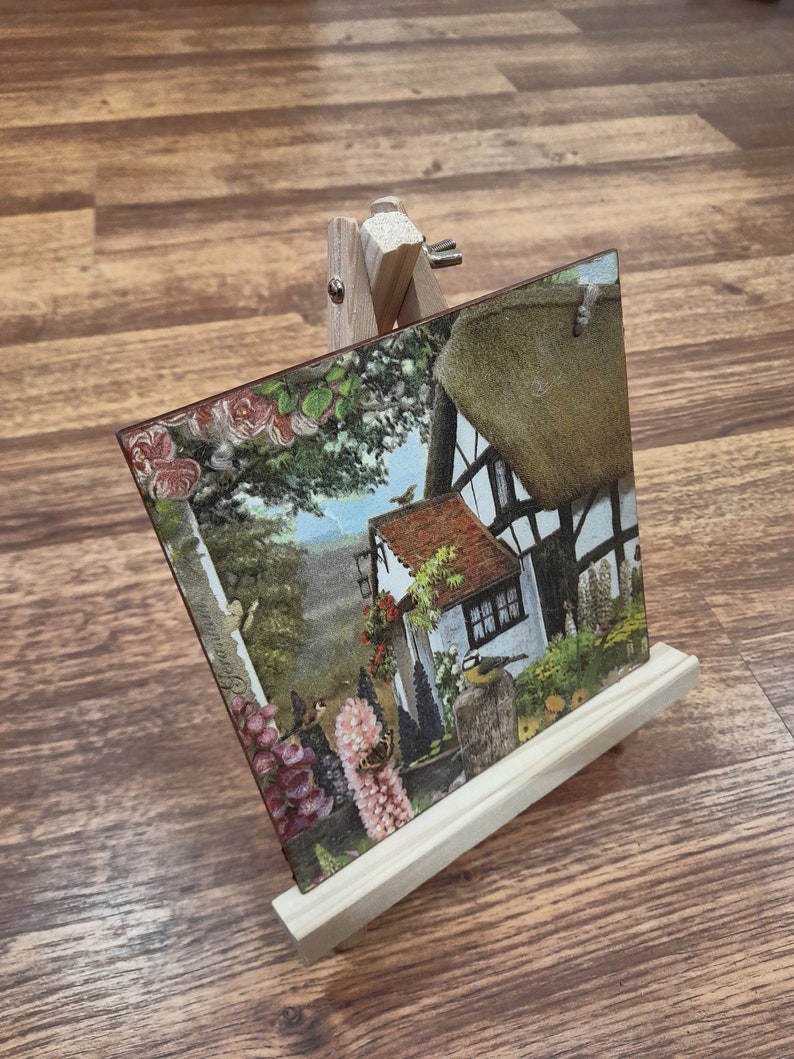 May include: A wooden easel with a small painting of a cottage with a thatched roof and flowers. The painting is on a wooden board and the easel is made of natural wood.