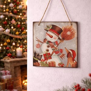 Peut inclure: Panneau carr&eacute; en bois de No&euml;l avec un bonhomme de neige en chapeau haut de forme et &eacute;charpe, tenant un balai. Deux chiens et des cadeaux sont &eacute;galement repr&eacute;sent&eacute;s. Le panneau est suspendu par une ficelle de jute.