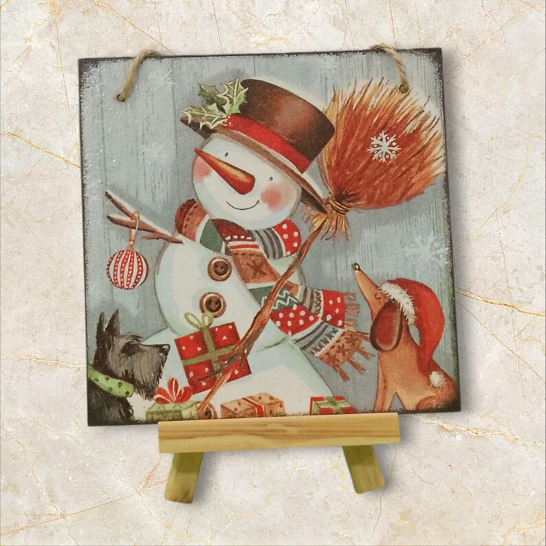 Peut inclure: D&eacute;coration de No&euml;l carr&eacute;e en bois repr&eacute;sentant un bonhomme de neige avec un nez en carotte, portant un chapeau et une &eacute;charpe. Un chien portant un bonnet de P&egrave;re No&euml;l et un autre chien sont &eacute;galement pr&eacute;sents. Le bonhomme de neige tient un balai et une boule de No&euml;l.