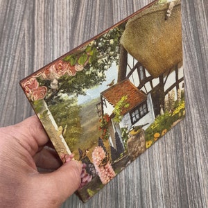 May include: A vintage-style postcard featuring a cottage with a thatched roof, surrounded by lush greenery and flowers. The postcard has a faded, antique look and includes a small bird in the upper right corner.