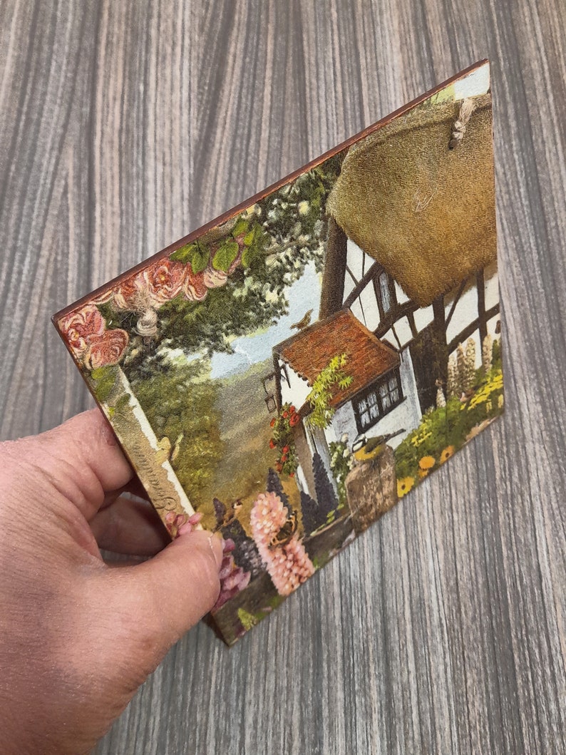 May include: A vintage-style postcard featuring a cottage with a thatched roof, surrounded by lush greenery and flowers. The postcard has a faded, antique look and includes a small bird in the upper right corner.