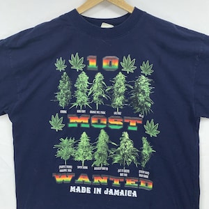 May include: A navy blue t-shirt featuring a graphic print with the text 10 MOST WANTED MADE IN JAMAICA. The design includes illustrations of ten green plant buds, small leaf icons, and the text labels Chronic, King Crypt, Orange Hill Magic, Cali Mist, AK-47, Purple Skunk, Super Skunk, Buskolo Shot-10, Get It Green, and Sticky Icky.