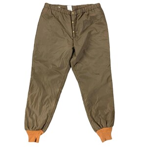 Op de afbeelding: Bruine geïsoleerde broek met elastische tailleband en knoopsluiting. De broek heeft oranje manchetten bij de enkels. Het materiaal heeft een lichte glans. Ontworpen voor koud weer.