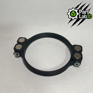 May include: A black circular object with two sets of silver magnets attached. The object has a matte finish and appears to be made of plastic. The logo "Part Lab3D" is visible in the upper right corner.